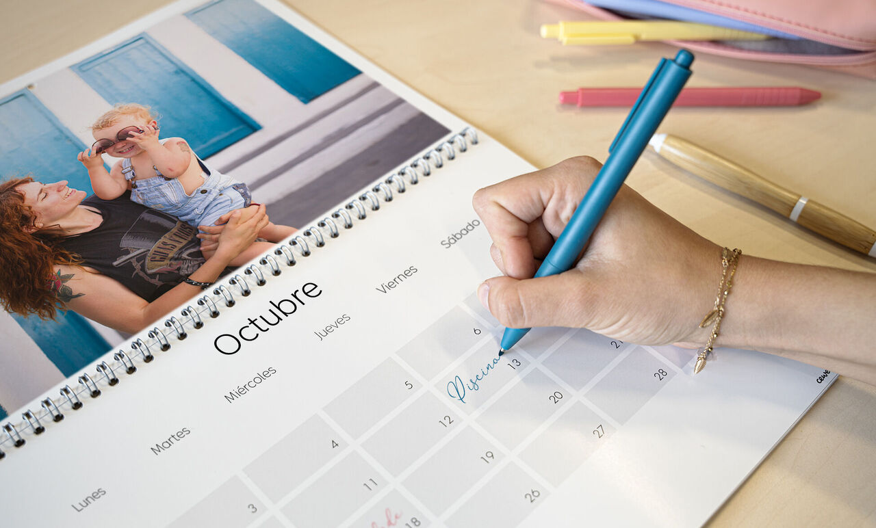 Una mano escribe en un colorido calendario de pared, mientras un niño con gafas de sol hace una foto.