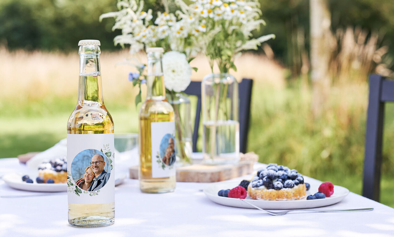 Dos botellas con etiquetas personalizadas sobre una mesa decorada, rodeadas de frutas y un pastel.