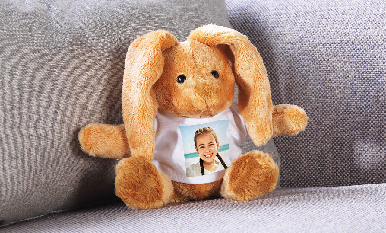 Un lindo conejo de peluche se sienta en un sofá. Lleva una camiseta con una foto.