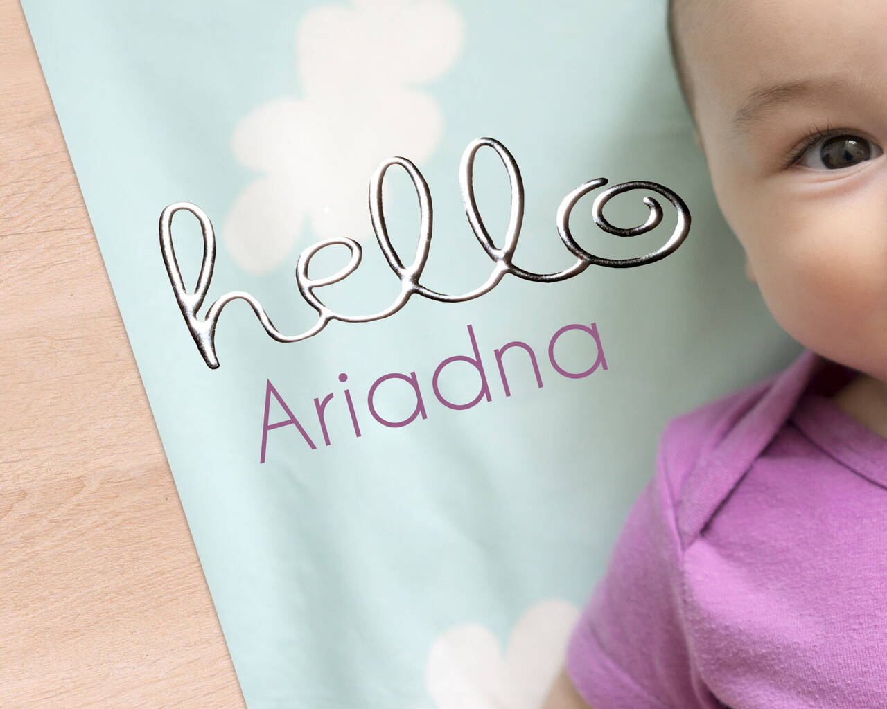 Un bebé sonriente tumbado sobre una manta azul clara con flores blancas y el texto "hello Ariadna".