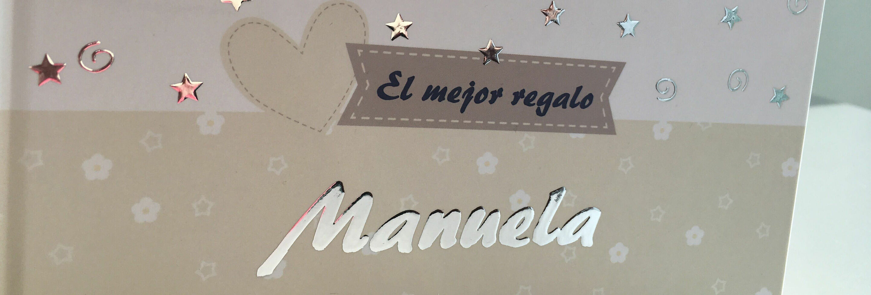 Un álbum CEWE titulado 'El mejor regalo' con el nombre 'Mannela' en una tipografía decorativa. La portada es elegante con estrellas brillantes y un diseño de corazón.