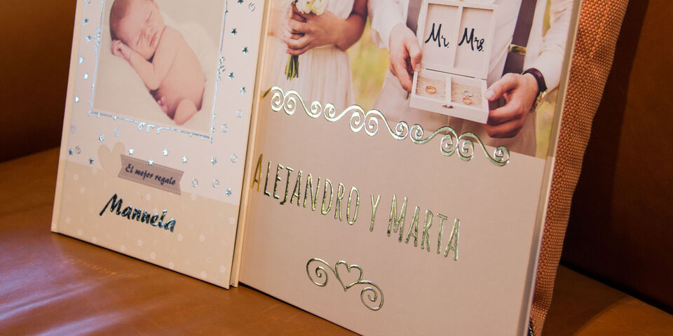 Dos libros de fotos bellamente diseñados, uno con una foto de bebé y el otro con los nombres Alejandro y Marta, decorados con brillo.