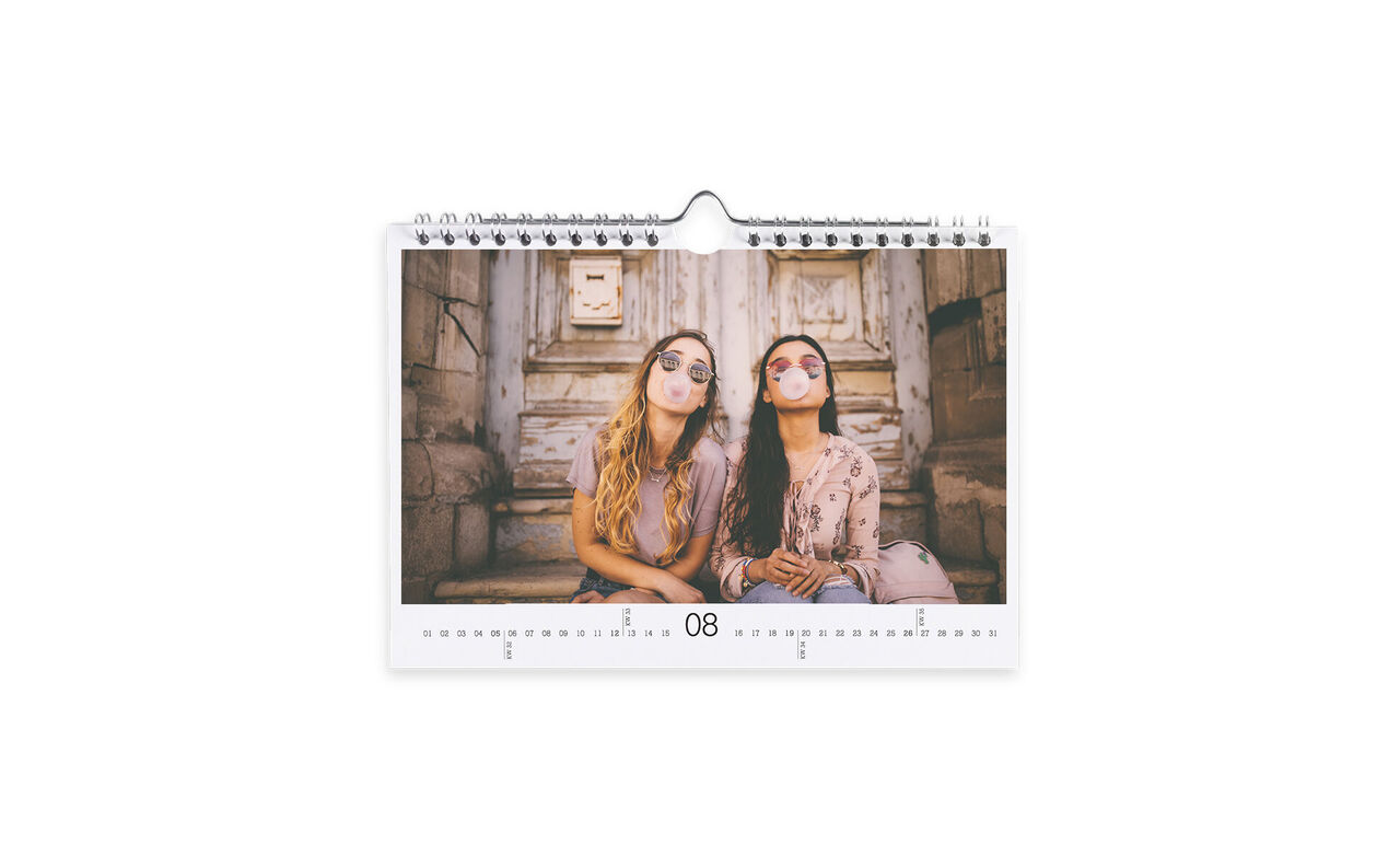 Dos mujeres sonríen frente a una pared rústica admirando un calendario de fotos CEWE