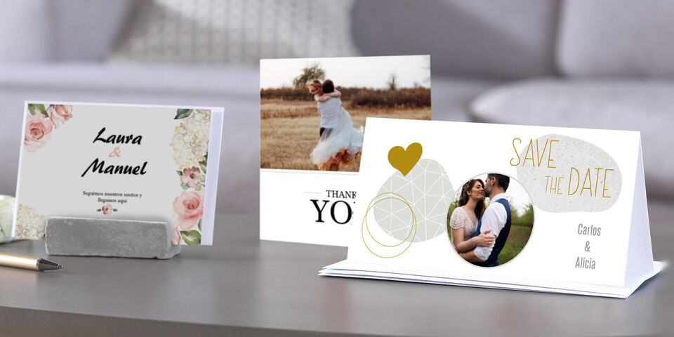 Tres tarjetas CEWE creativas sobre una mesa, con imágenes alegres y textos. Una presenta una pareja, otra dice 'GRACIAS' y la tercera dice 'RESERVA LA FECHA'.