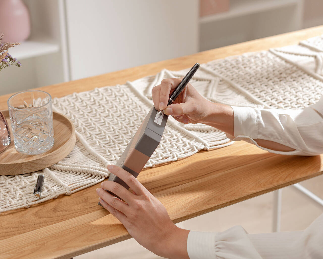 Una persona sostiene un cuaderno gris y escribe con un bolígrafo negro. La mesa es de madera.
