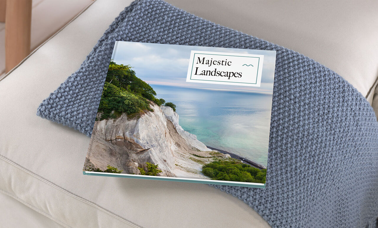 Un álbum de fotos CEWE abierto titulado "Majestic Landscapes" sobre un sofá beige, rodeado por un cojín azul.